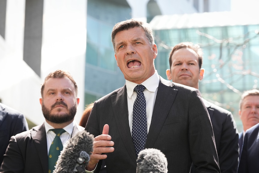 A man in a suit and tie talks animatedly into microphones, with other men standing behind him.