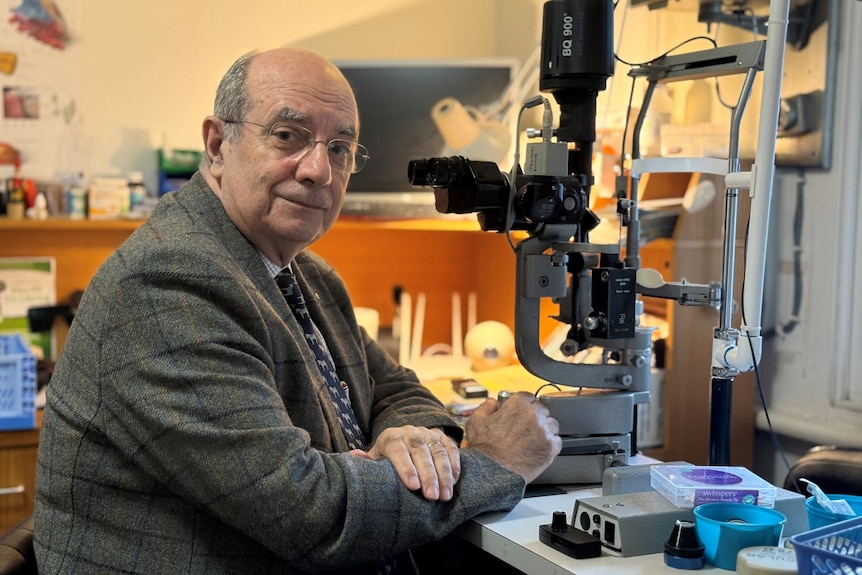 An older man wearing glasses and a smart jacket sits at a desk with ocular equipment on it.