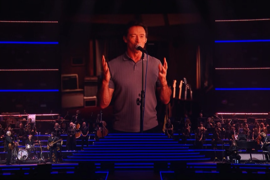 Hugh Jackman on a screen. He raises his hands and speaks into a mic.