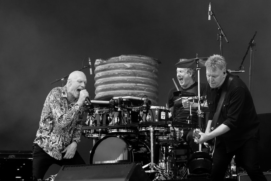 Peter Garrett sings into a microphone. His bandmates nearby are on guitar and drums.
