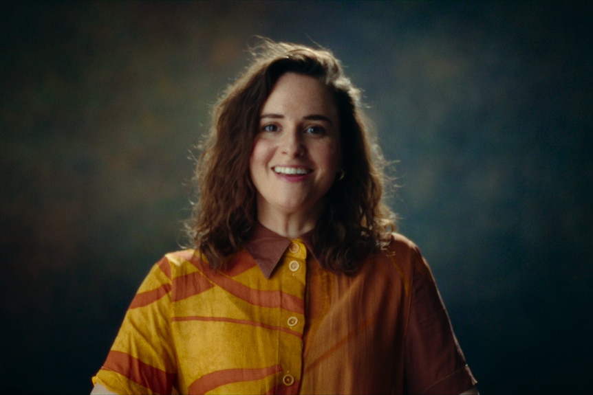 A white woman with long brown hair, smiling and wearing a yellow and orange top