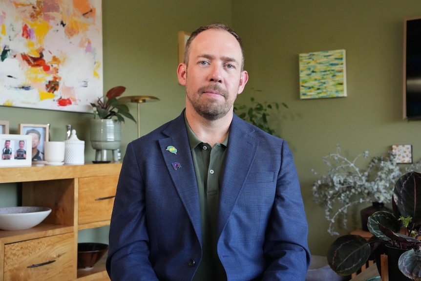 A bearded man wearing a blue jacket sits in a green-roomed wall with a serious expression on his face.