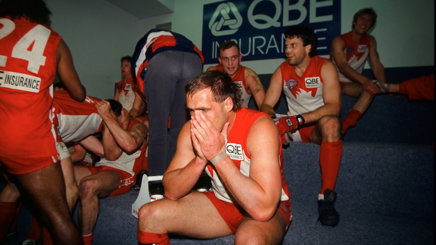 Sydney Swans star Tony Lockett has his hands over his mouth as he sits in a room with celebrating teammates after a big win.