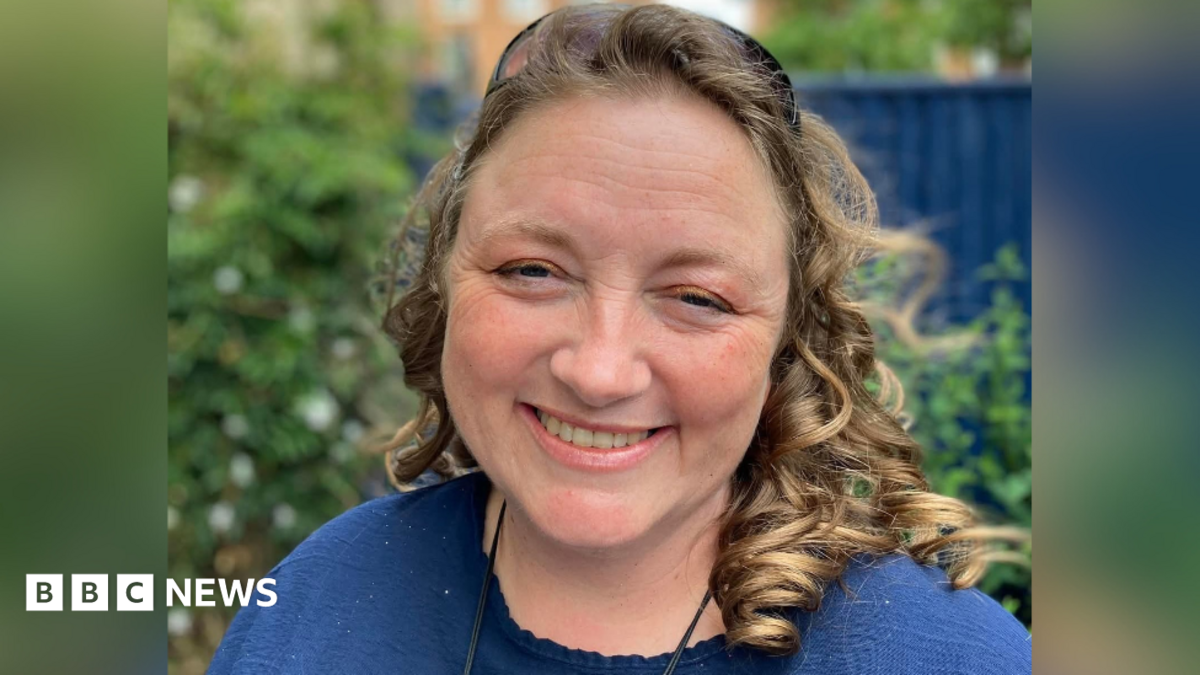 A woman with curled hair smiling into the camera. She is wearing a blue top and is stood in a garden, with the background slightly blurred.