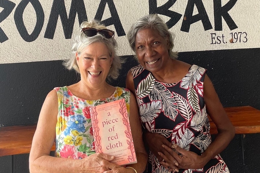 Two woman stand side by side and hold a book with a red cover