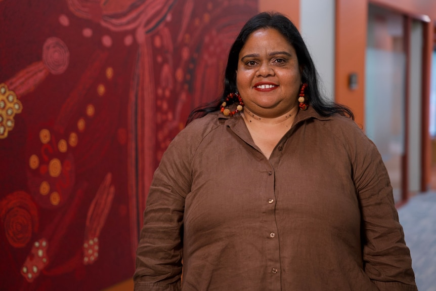 Sam Murray wears a brown shirt. She has long hair and beaded earrings and is standing in front of an Indigenous painting.
