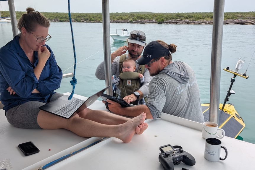 Fisherman Tiger Davey and family members on a boat in water, they are looking at laptop and tablet screens with worried faces.