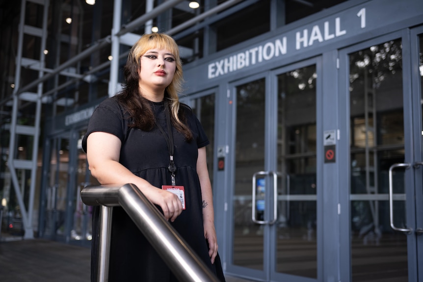 A young woman with black-and-tan hair wears a dark dress while standing near the entry to a large venue.