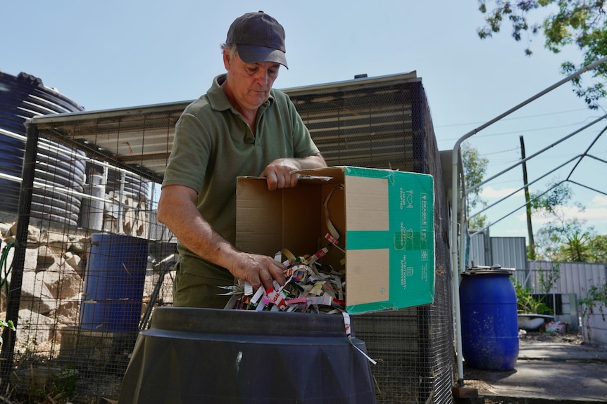 A man wearing a green T-shirt and cap empties shreds of papers from a cardboard box into a black bin.