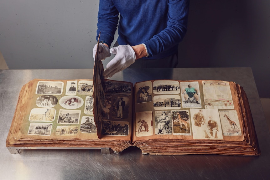 A person wearing a blue top and white cotton gloves turns the pages of an old scrapbook full of photos.