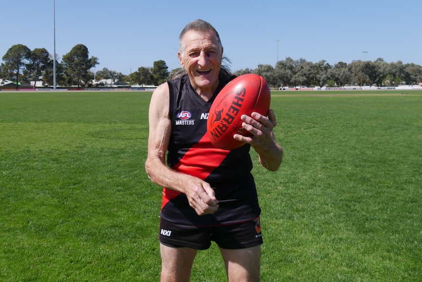 An elderly man in a black and red football guernsey handballs a red Australian Rules football.