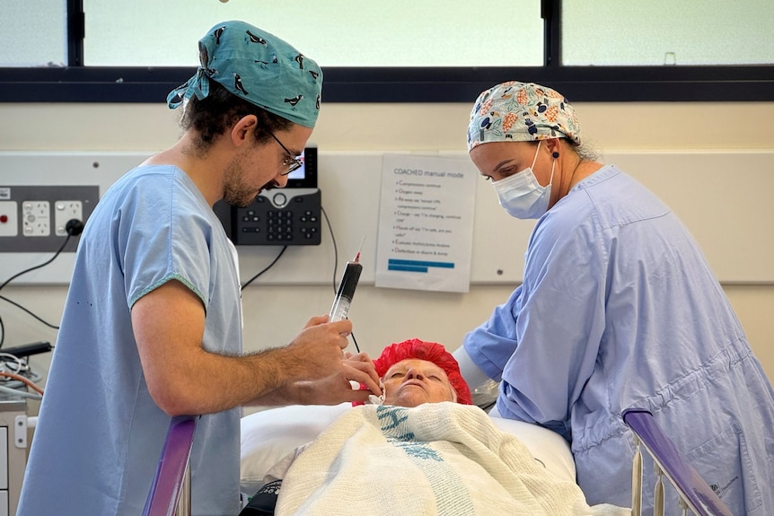 Two nurses in blue scrubs stand on either side of a hospital bed, with one holding a huge needle above a patient. 