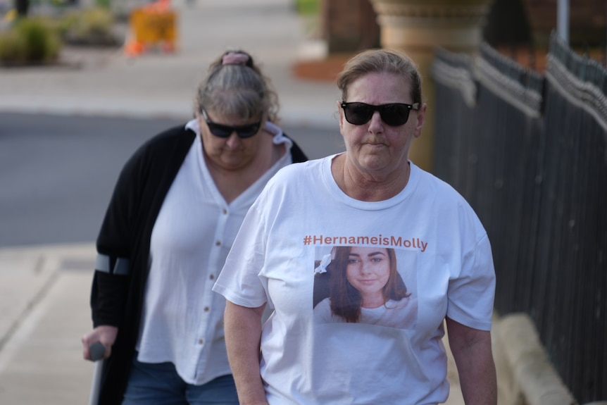 A woman, in a white shirt with a photo of a woman and words on it, walking on the footpath.