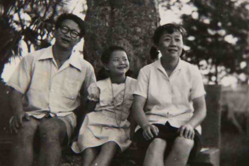 A black and white image of a little girl in a dress seated on a bench next to her mum and dad outside.