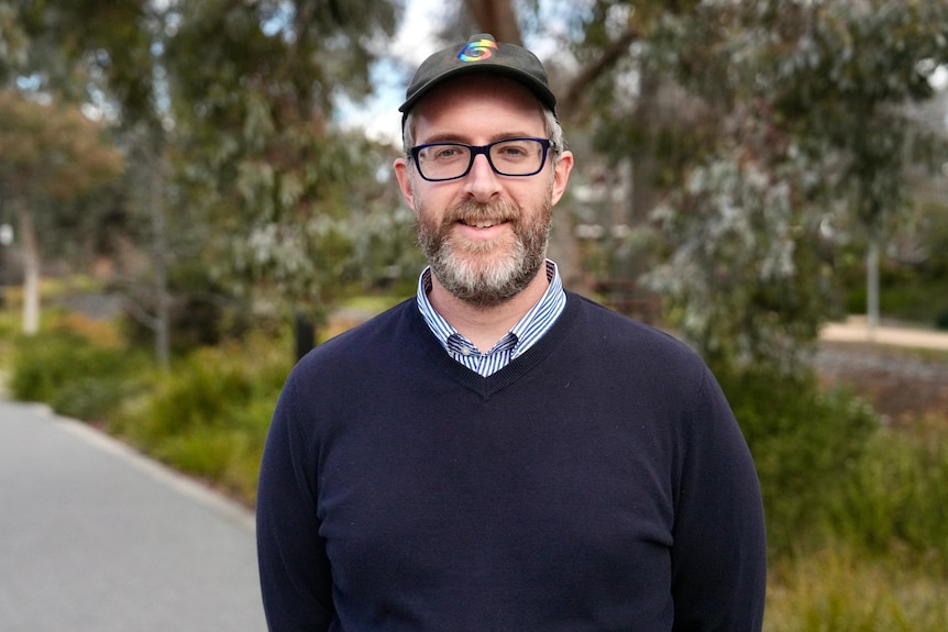 A white man with short beard and cap and glasses smiles with his hands behind his back.
