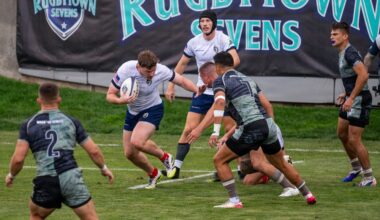 A rugby sevens match is in progress at the Rugbytown Sevens tournament, with players in white uniforms (appearing to represent the USA based on the flag patch) competing against players in dark camouflage-patterned jerseys. The ball carrier in white is attempting to evade two defenders while a teammate in a scrum cap provides support from behind. The action takes place on a well-maintained grass field in front of prominent tournament branding banners.