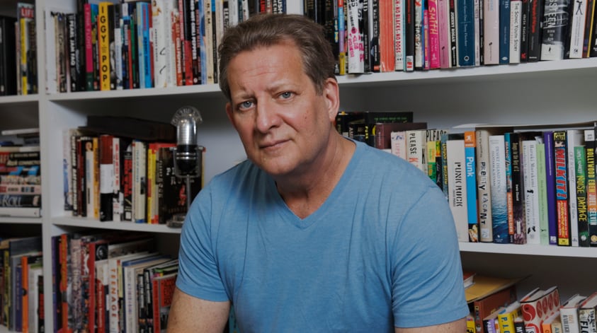 A man with brown hair and a blue t-shirt sits in front of a bookcase.
