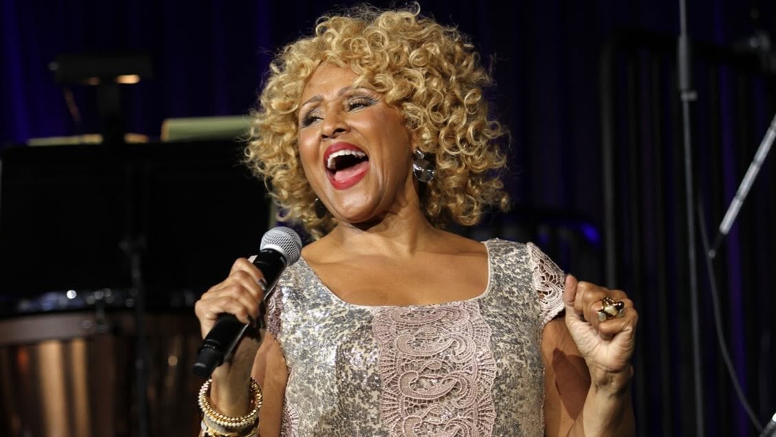 A Black woman with curly blonde hair holds a microphone and sings.