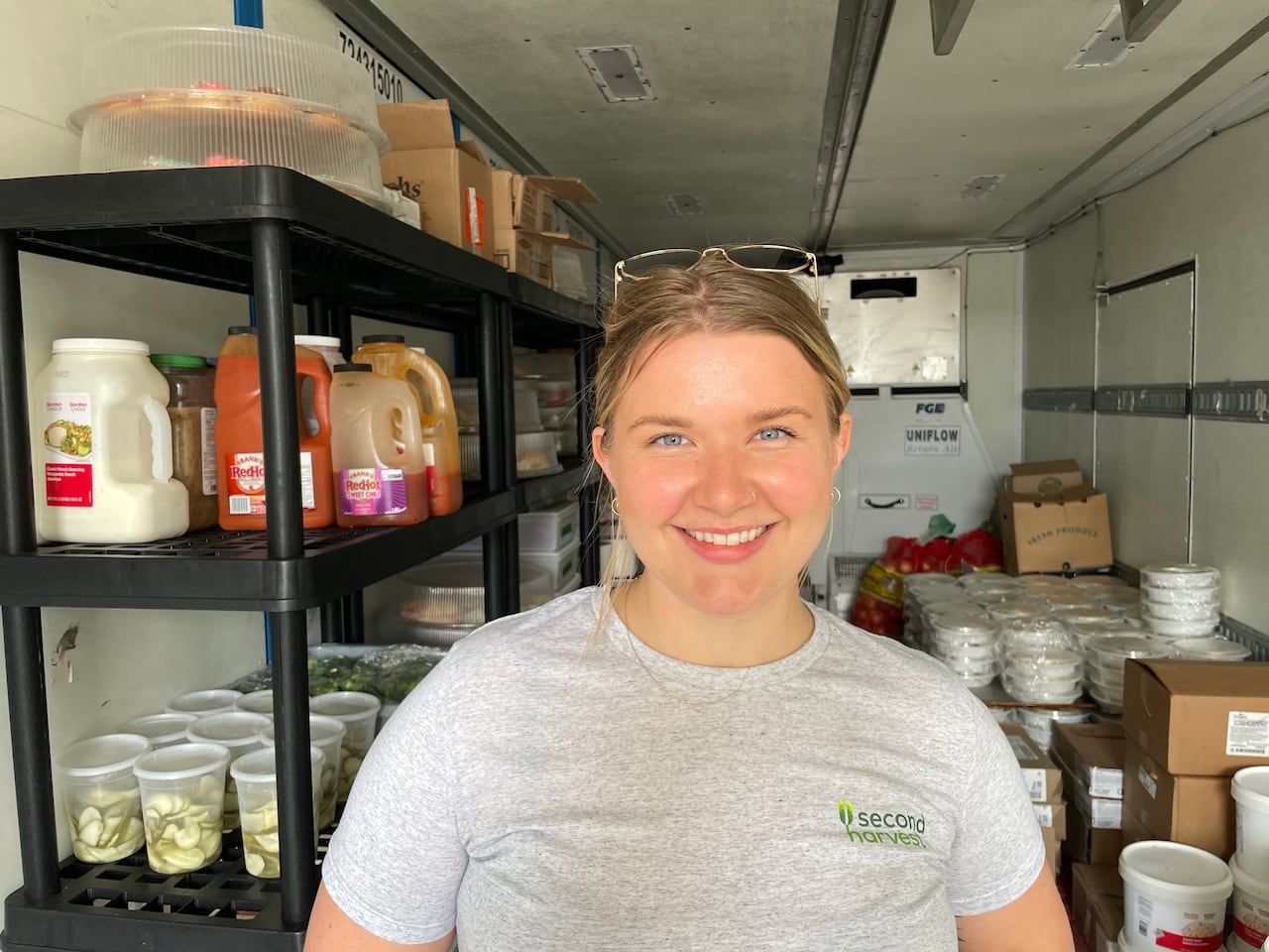 A woman in a gray t-shirt stands in the back of a cooled truck, behind her food and food products in packages and boxes can be seen.