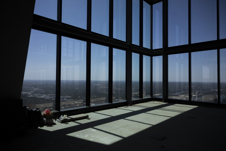 A sunlit, empty room with floor-to-ceiling windows offers a panoramic view of a urban landscape and hill country.