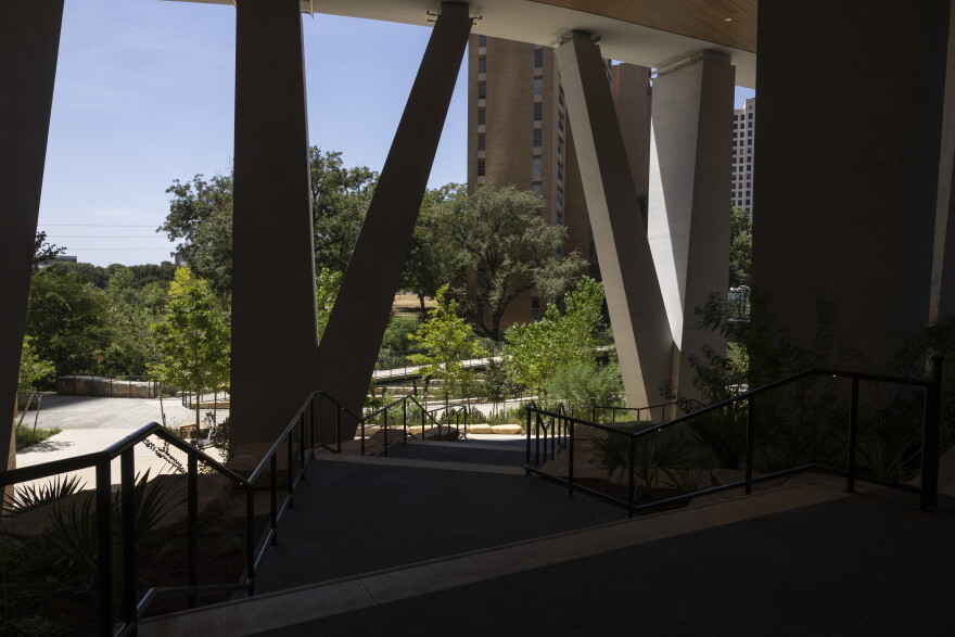 A modern architectural structure with bold support columns frames a sunlit outdoor scene featuring trees and a paved path.