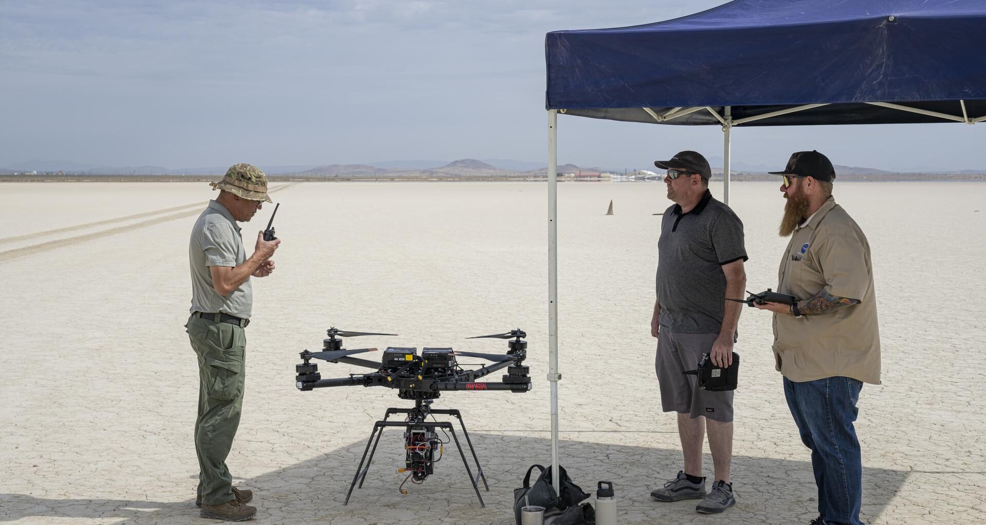 One man communicates with a hand-held radio, as two other men wait to the side for the go-ahead command to fly an experiment on a drone.