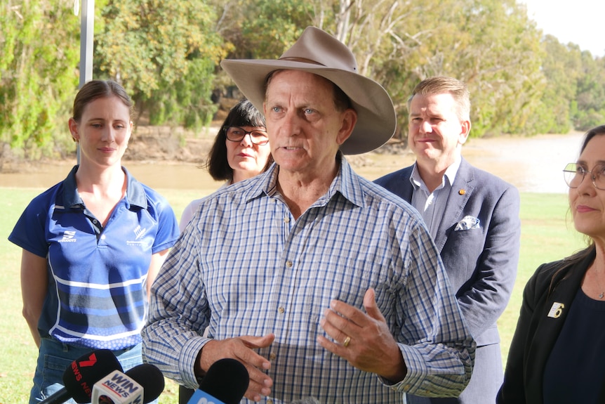 A man in an Akubra hat speaks passionately at a press conference, flanked by four other people in front of a river.