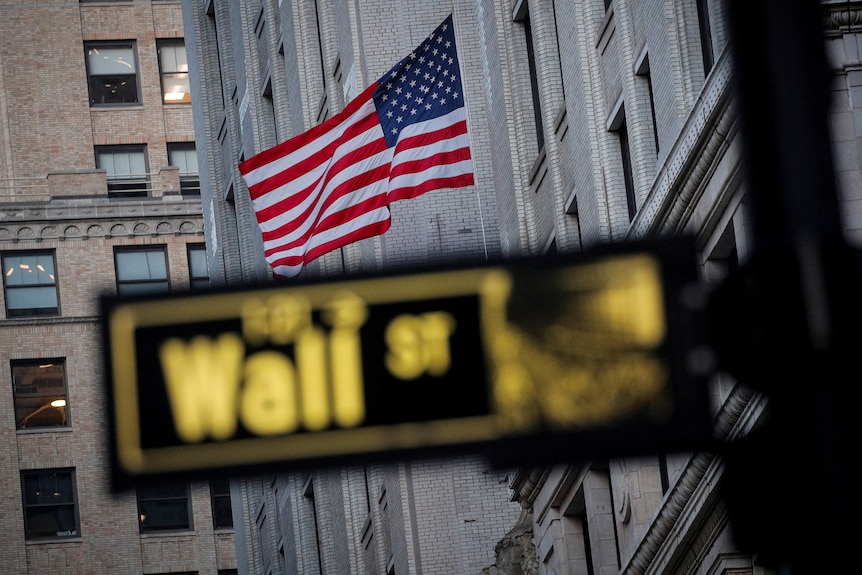 The US flag is flying on the side of a building with a blurred street sign for Wall St in the foreground.