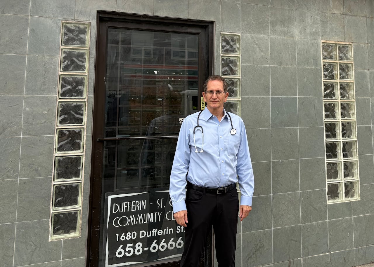A man in a blue button-up shirt and black pants with a stethoscope around his neck stands outside a clinic.