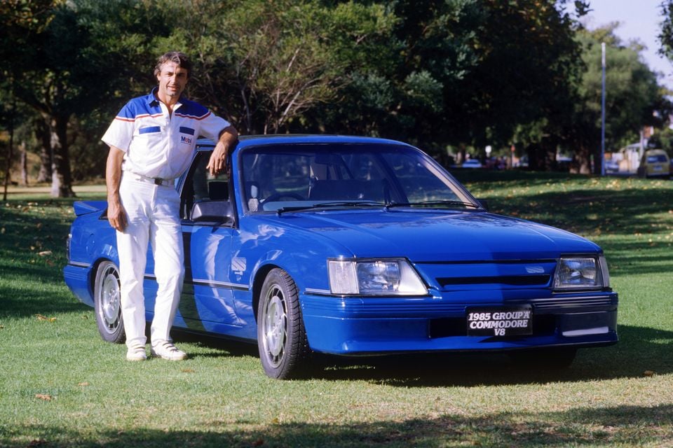A man in a Mobil-branded racing outfit stands beside a blue 1985 Holden Commodore VK Group A V8 sedan on a grassed area outdoors.
