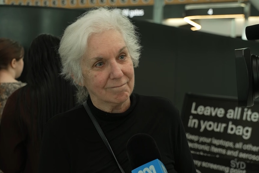Sue Hobley smiling, speaking to a camera, with a blue ABC microphone to the side. She has white hair and wears a black jumper.