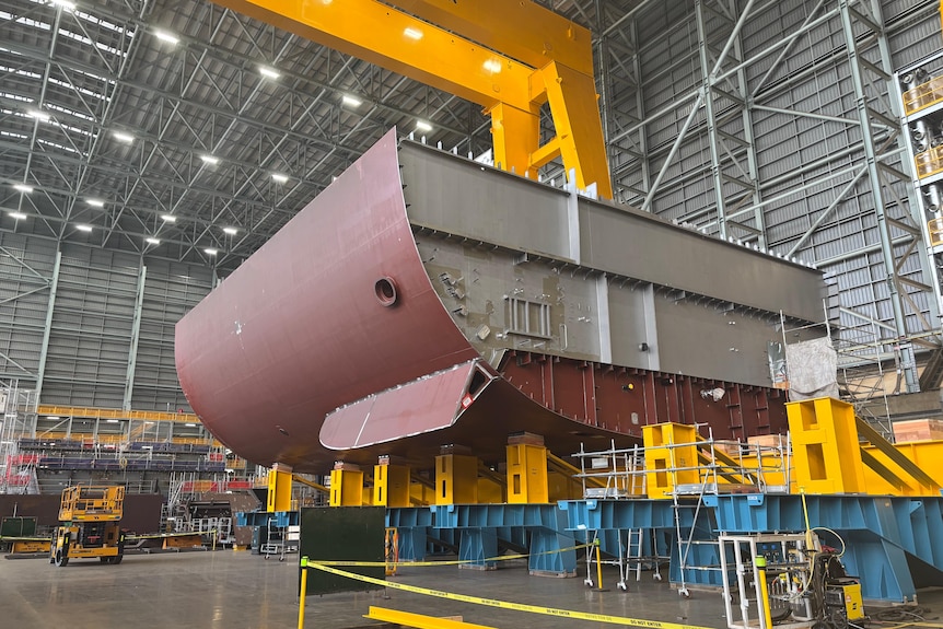 A part of a large ship being put together in a large warehouse site.