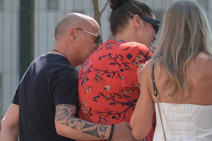 the back of a group of three people hugging and crying at the scene of a crash where an officer died