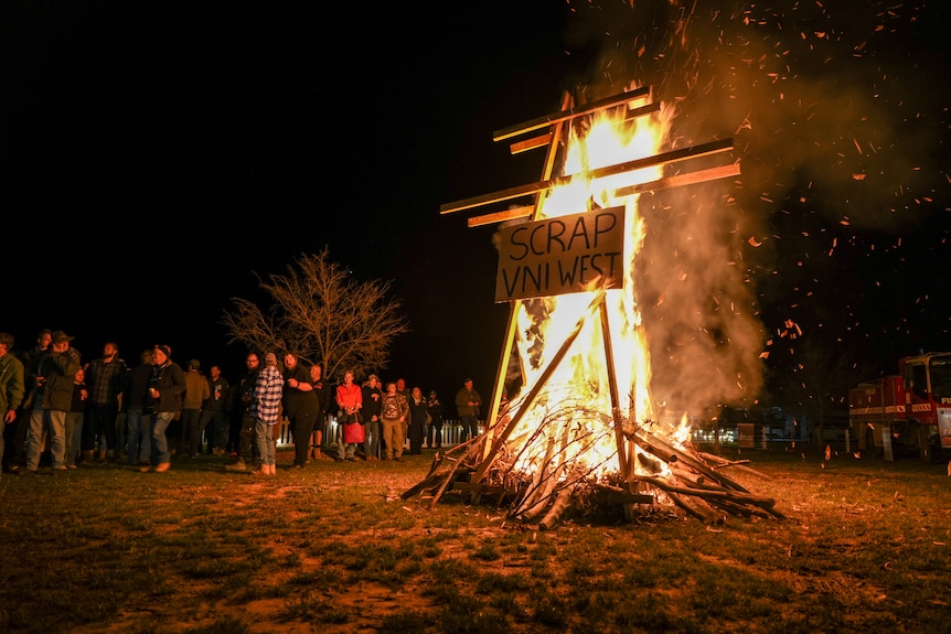 Crowd of farmers surround a fire, where a wooden prop transmission line and a sign reading 'SCRAP VNI WEST' are burning