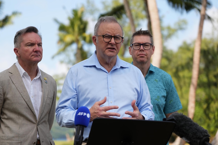 The Prime Minister in blue button-down shirt speaks flanked by two other ministers with palm trees in background