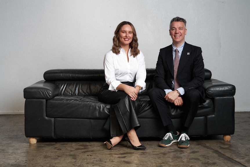 a man and a woman sit on a couch in business attire, smiling to the camera