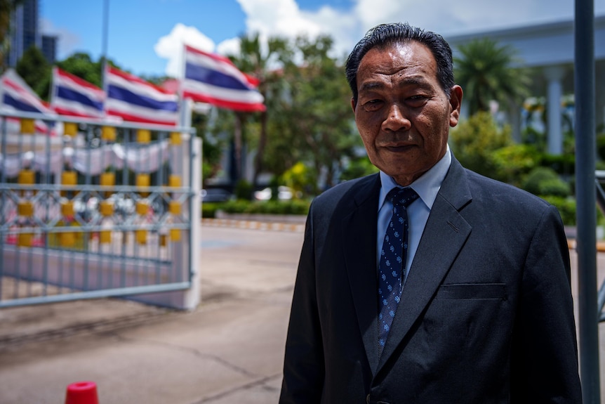 A man wearing a blue suit standing outside a gate adorned with Thai flags.