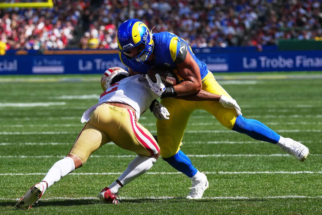 A Los Angeles Rams offensive player wearing a blue and yellow uniform tightly clutches the football while leaning into a tackle from a San Francisco 49ers defender clad in a white shirt and gold pants.