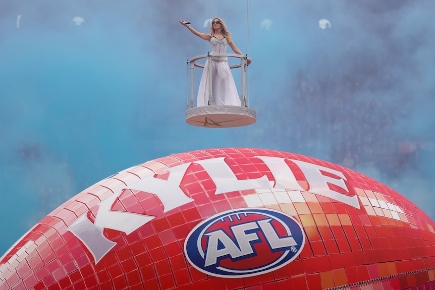 Kylie Minogue singing in a floating platform above a massive red AFL football that says "Kylie"