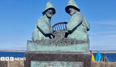 The photo shows the completed St Ives Mariners Memorial on a pier overlooking the sea. The bronze sculpture depicts two fishermen sitting opposite each other working with fishing nets between them. They are wearing oilskins.