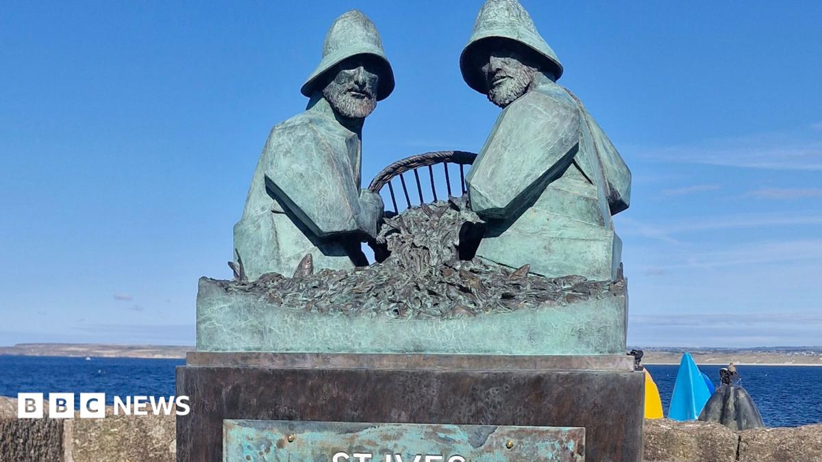 The photo shows the completed St Ives Mariners Memorial on a pier overlooking the sea. The bronze sculpture depicts two fishermen sitting opposite each other working with fishing nets between them. They are wearing oilskins.