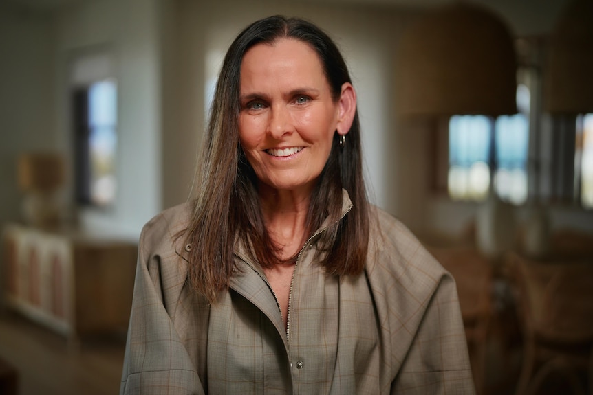 A woman with long brown hair poses for a photo inside, looking into camera and smiling.