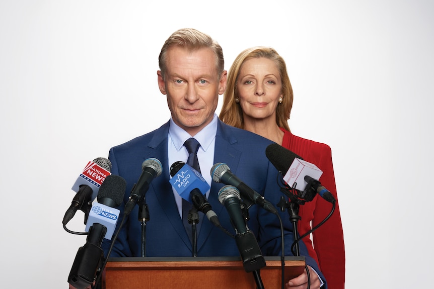 A suited Richard Roxburgh, 64, stands behind a lectern, looking determined, Heather Mitchell, 68, standing behind him.
