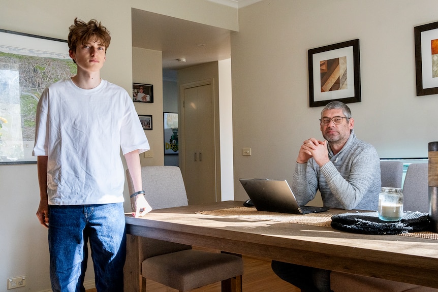 A 16-year-old boy stands at a desk where his father is sitting.