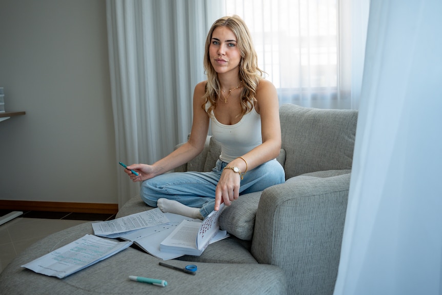 A young woman sits on a grey couch with study books in front of her.