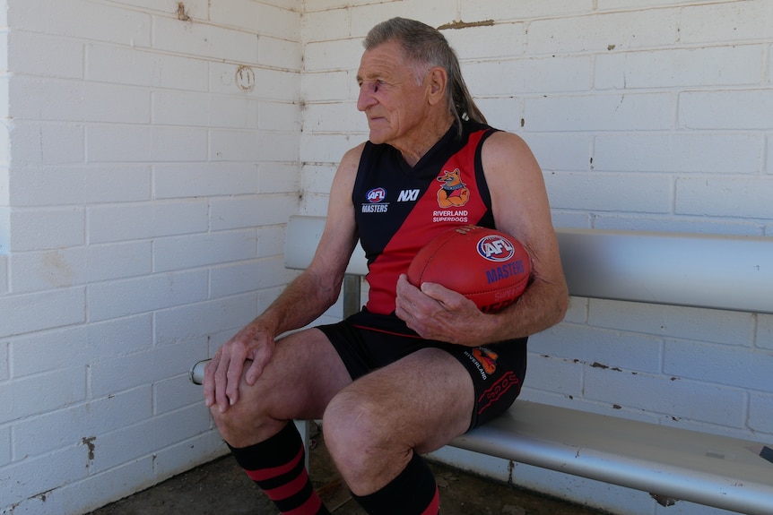 An elderly man in a football guernsey holds a red Australian Rules football while sitting on the bench.