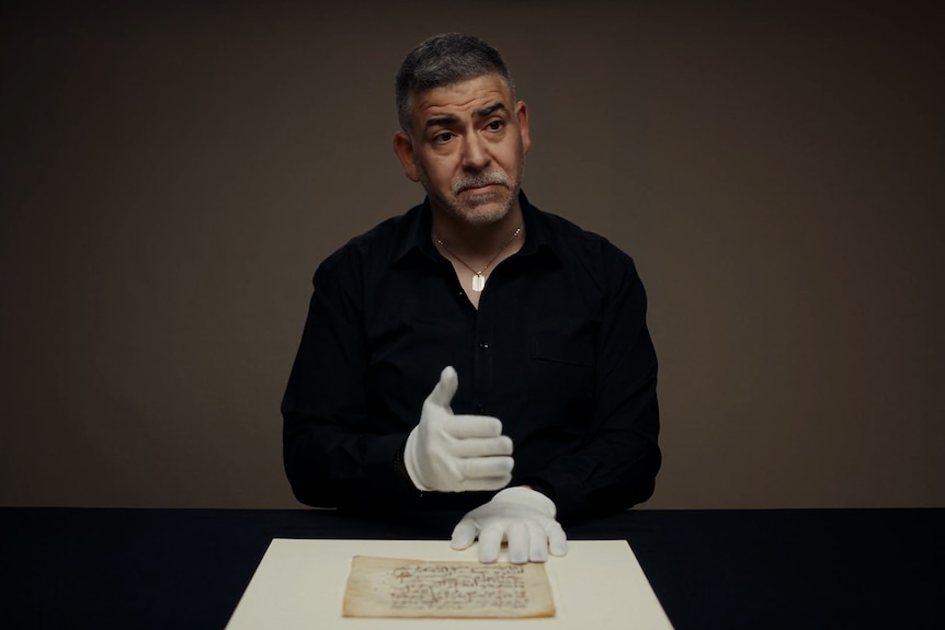 A greying dark-haired man with a short beard wearing white gloves handling a manuscript.