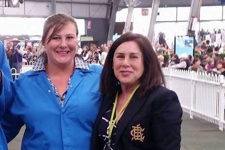 A smiling lady (Sapphire Halliday) in a blue coat and an older lady (Deborah Halliday) pose in a cattle arena.