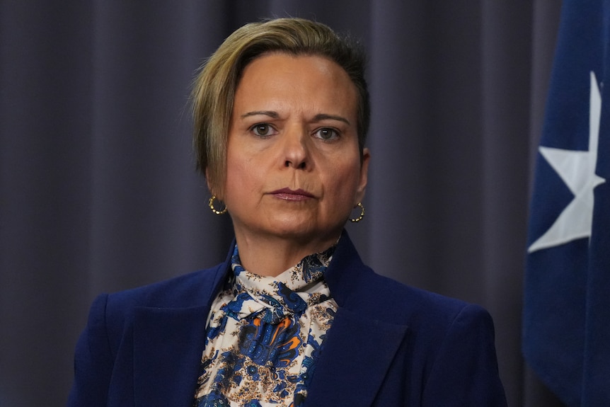 A woman in a blue suit and floral shirt looks sternly at camera.