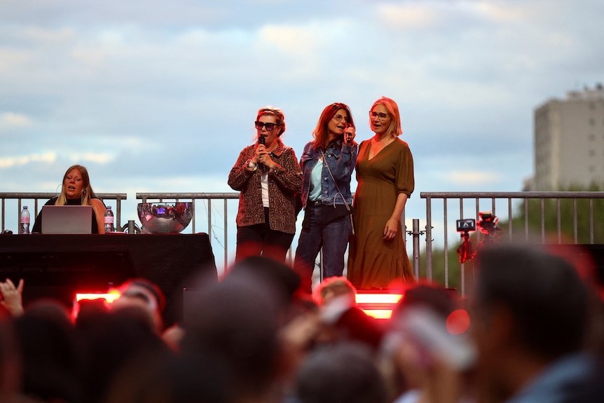 Three women clutch a microphone on stage with red lights flashing in the crowd.